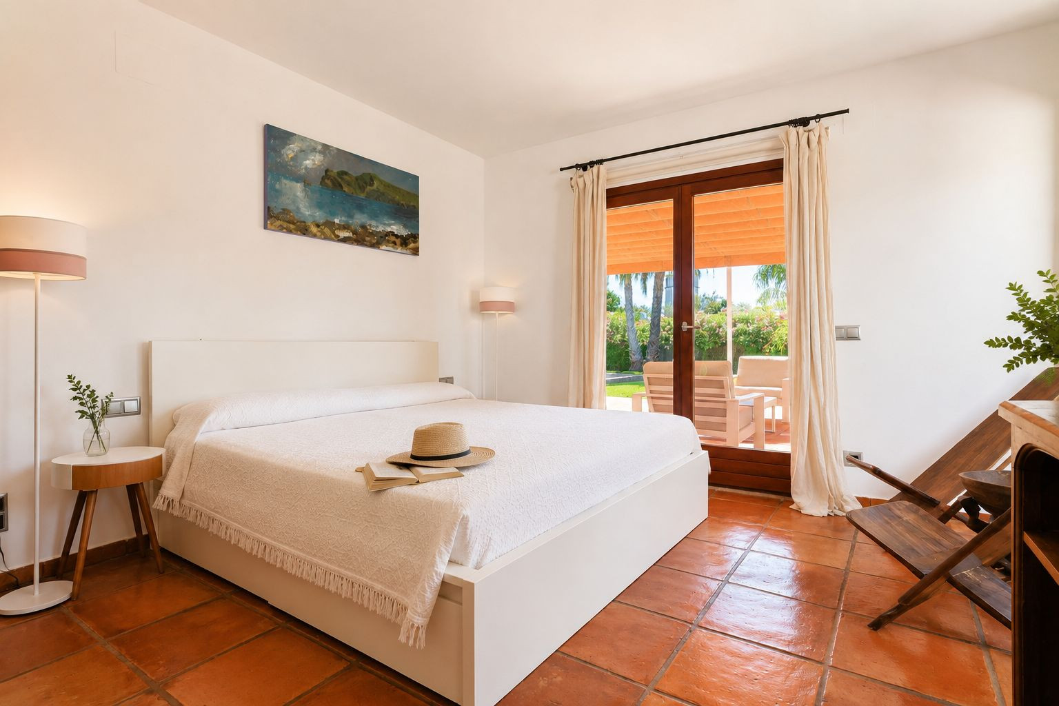 Bright bedroom featuring a white bed, artisanal terracotta floors, and direct access to an outdoor terrace with garden views and seating.