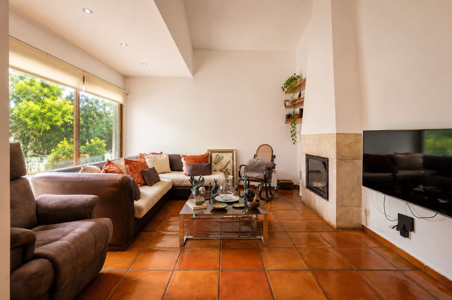 Bright living room featuring traditional terracotta floors, a stone-clad fireplace, and large windows overlooking lush greenery.