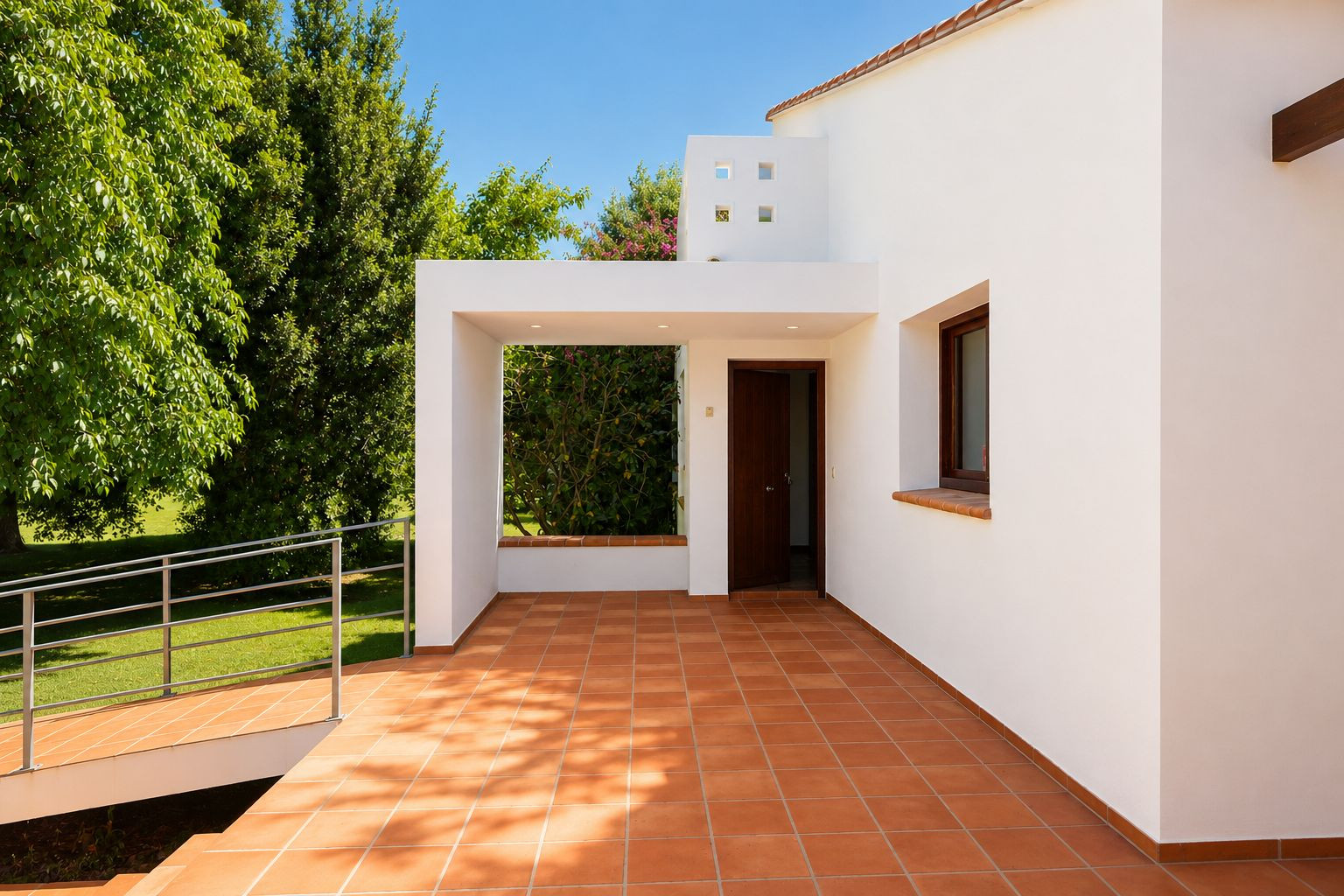 Covered main entrance patio featuring terracotta floor tiles and recessed lighting. An accessible ramp leads to the lush garden, complementing the white facade and dark wood door.