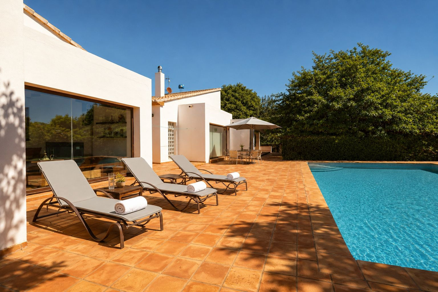 Expansive terracotta-tiled pool terrace featuring three sun loungers, a shaded outdoor dining area, and a clear blue swimming pool. The white facade with large windows reflects the surrounding greenery.