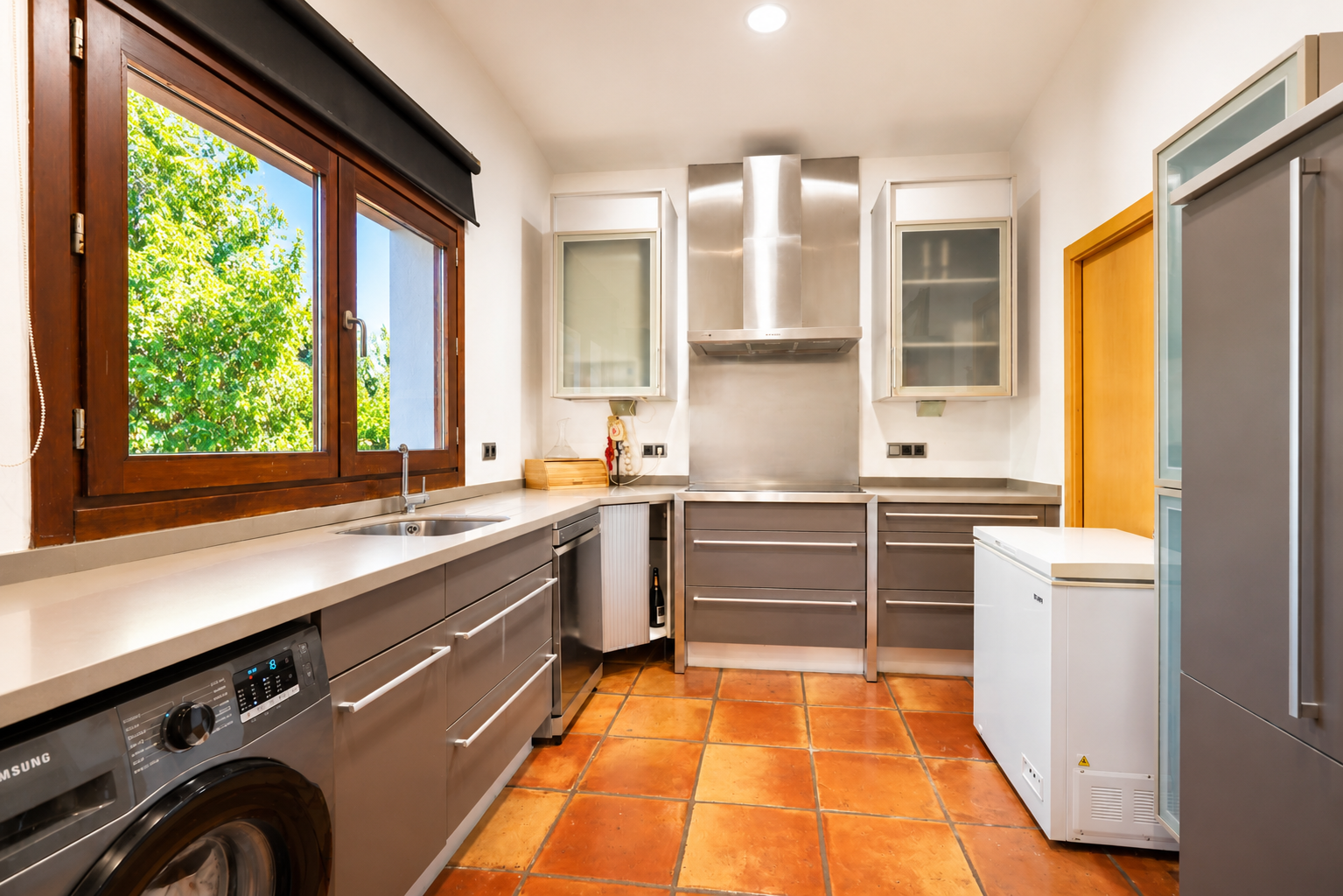 Functional kitchen featuring an integrated washing machine, artisanal terracotta floor, and a large wooden window overlooking the garden.
