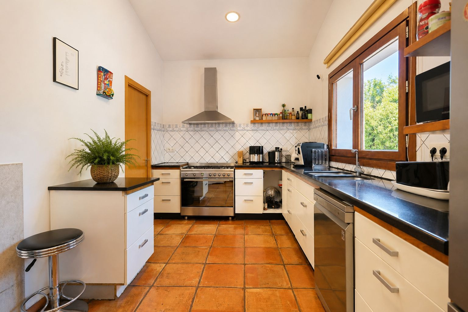 Functional kitchen featuring artisanal terracotta floors, dark countertops, and large wooden-framed windows providing abundant natural light.
