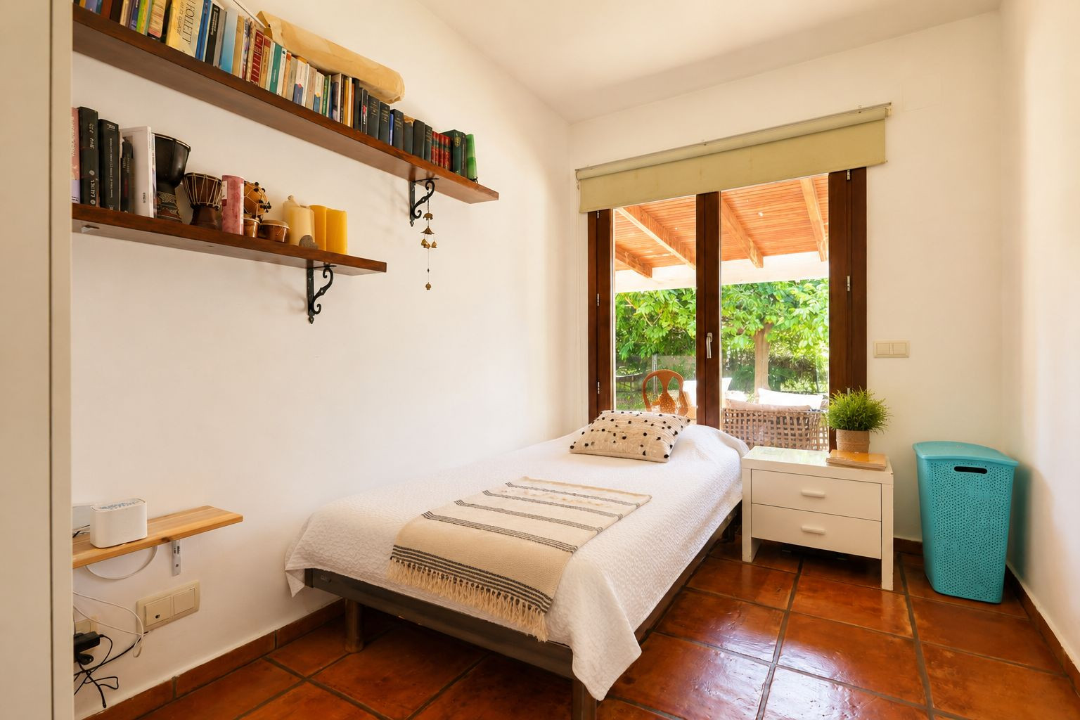 Individual bedroom featuring terracotta tiled floors, white walls, and direct access to a covered terrace through a large wooden-framed glass door. Includes wall-mounted shelves with books.