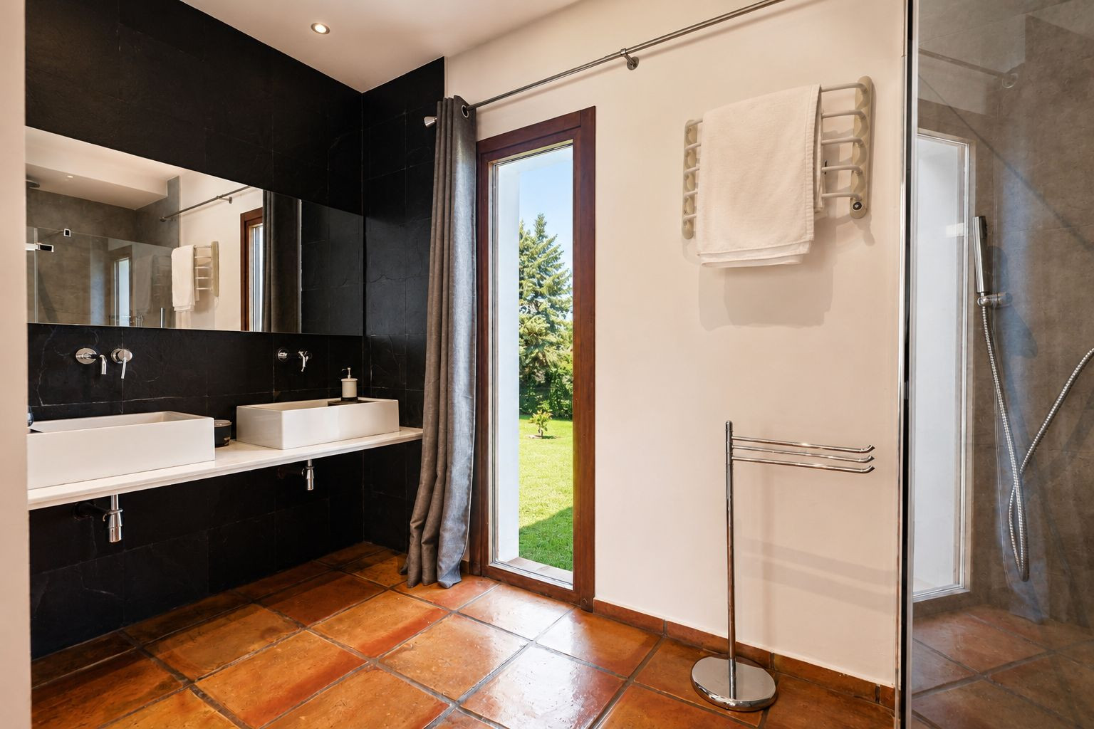 Spacious bathroom featuring terracotta floor tiles, a double vanity with rectangular white sinks against a black tiled wall, and a large window offering garden views. Includes a glass-enclosed shower.