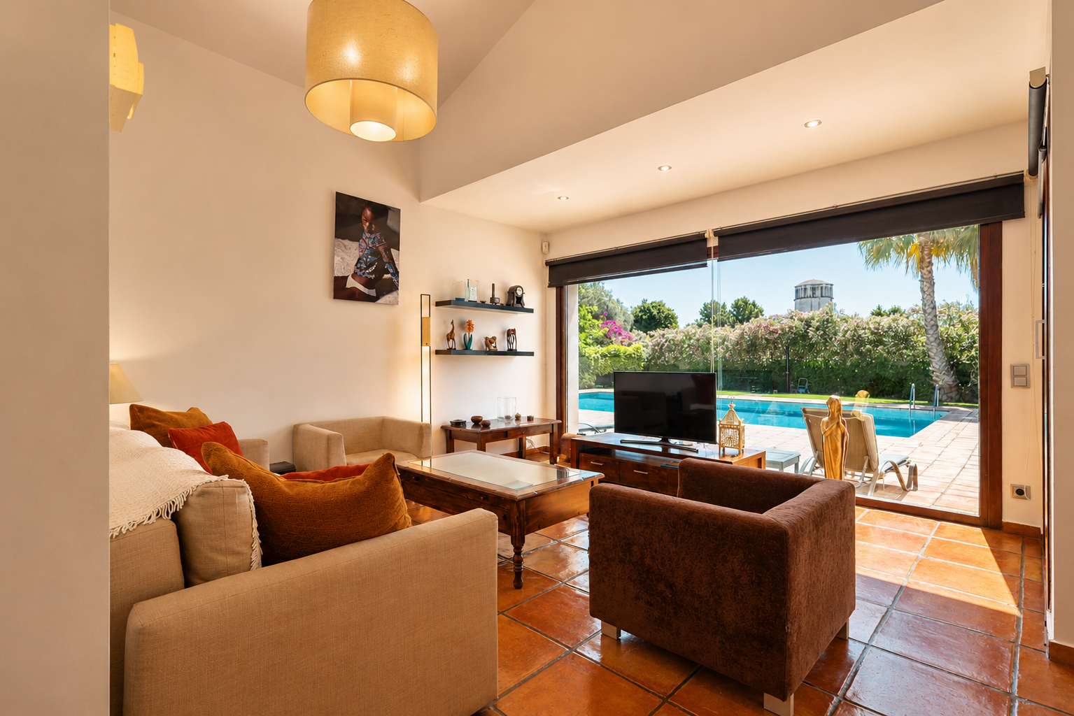 Spacious living room featuring terracotta tile floors and direct access to the swimming pool. Natural light fills the room, offering comfortable seating and garden views with a distant tower.