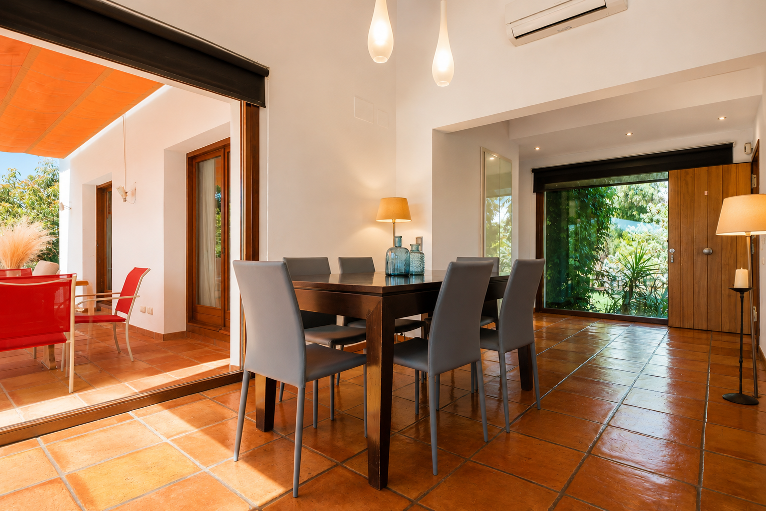 The bright dining area features a dark wood table with six grey chairs, set on terracotta floors. Large glass doors open to an outdoor terrace with an orange awning, while another window offers lush garden views.