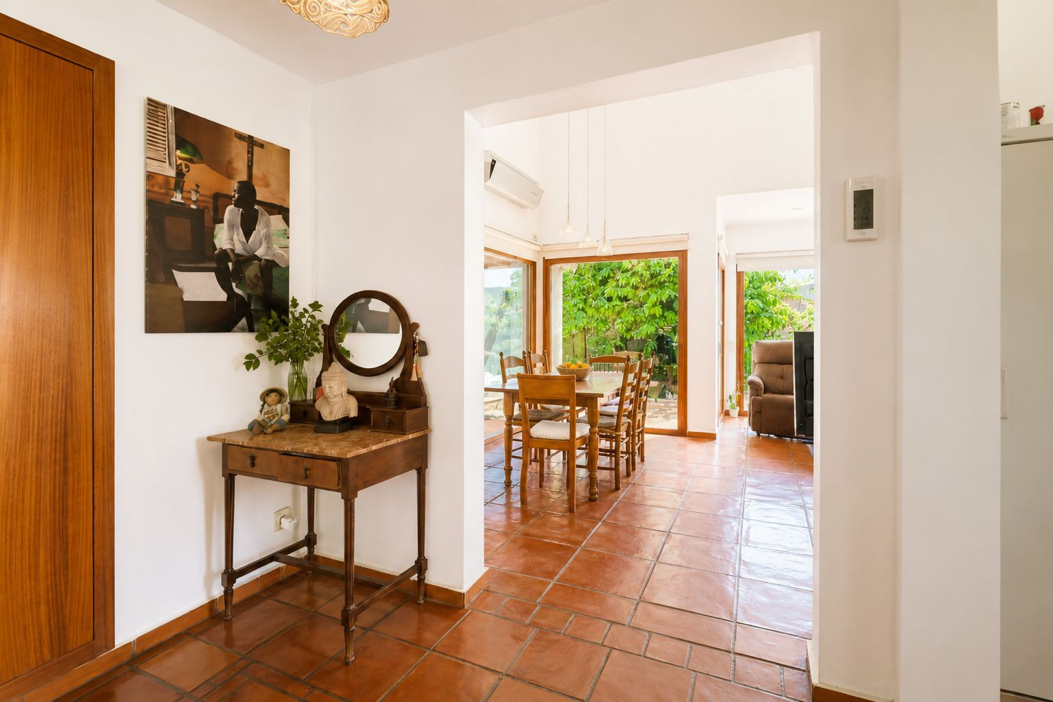 The bright entryway features traditional terracotta flooring and a vintage console table with mirror, opening into a spacious dining area with large windows overlooking lush garden views.