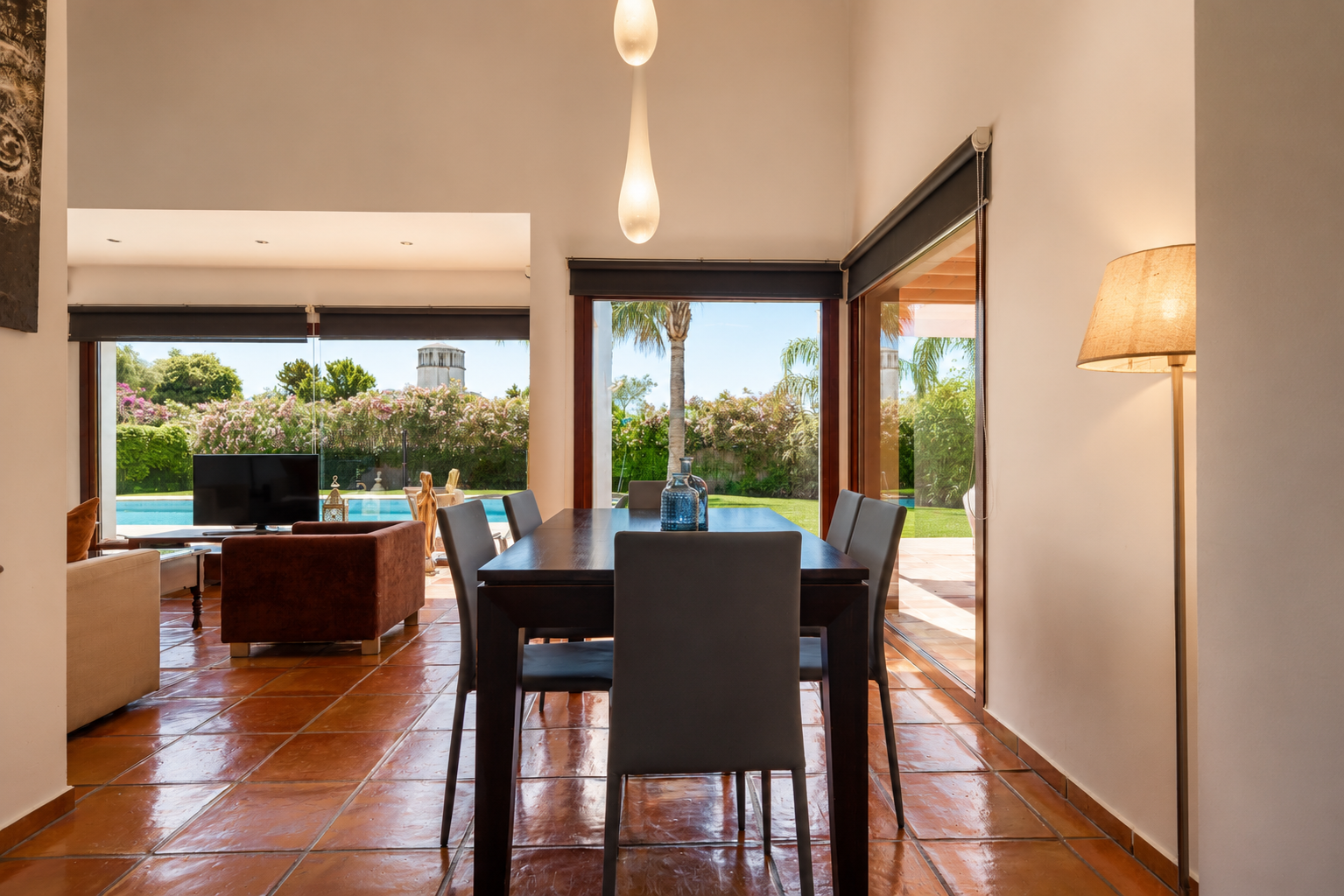 The dining area features a dark wooden table and terracotta floors, illuminated by natural light from large glass doors. It offers direct views of the lush garden, swimming pool, and a distant tower.