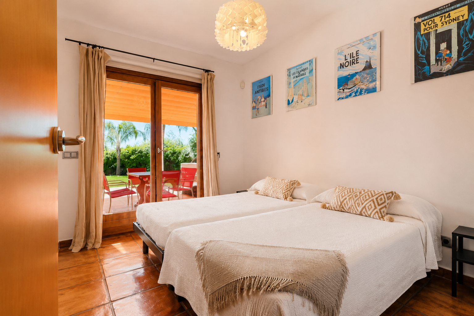 Twin bedroom featuring terracotta floors and direct access to a private outdoor terrace. The large glass door reveals a garden with a palm tree and red seating.