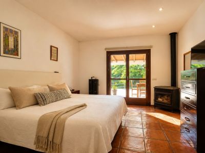 Bright bedroom featuring a large bed, traditional terracotta floor tiles, and a cozy wood-burning stove. Double glass doors open to a private outdoor terrace amidst lush greenery.