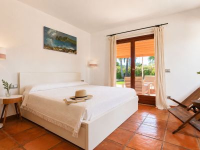 Bright bedroom featuring a white bed, artisanal terracotta floors, and direct access to an outdoor terrace with garden views and seating.