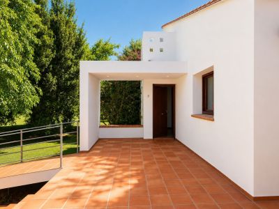 Covered main entrance patio featuring terracotta floor tiles and recessed lighting. An accessible ramp leads to the lush garden, complementing the white facade and dark wood door.