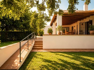 Covered naya with exposed wooden beams and an outdoor dining area, overlooking a manicured lawn and lush Mediterranean garden, accessible via tiled ramp and steps.