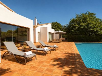 Expansive terracotta-tiled pool terrace featuring three sun loungers, a shaded outdoor dining area, and a clear blue swimming pool. The white facade with large windows reflects the surrounding greenery.