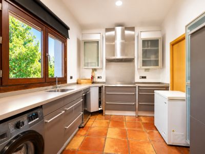 Functional kitchen featuring an integrated washing machine, artisanal terracotta floor, and a large wooden window overlooking the garden.