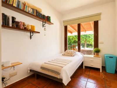 Individual bedroom featuring terracotta tiled floors, white walls, and direct access to a covered terrace through a large wooden-framed glass door. Includes wall-mounted shelves with books.