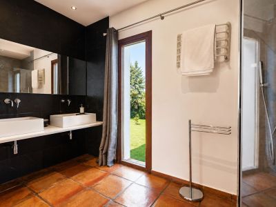 Spacious bathroom featuring terracotta floor tiles, a double vanity with rectangular white sinks against a black tiled wall, and a large window offering garden views. Includes a glass-enclosed shower.