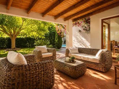 Spacious covered terrace with a wooden beamed ceiling and terracotta floor, featuring woven outdoor seating and direct access to the lush garden and pool area.