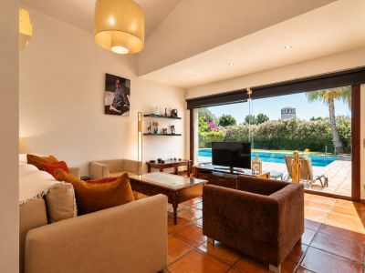 Spacious living room featuring terracotta tile floors and direct access to the swimming pool. Natural light fills the room, offering comfortable seating and garden views with a distant tower.