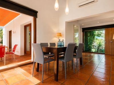 The bright dining area features a dark wood table with six grey chairs, set on terracotta floors. Large glass doors open to an outdoor terrace with an orange awning, while another window offers lush garden views.