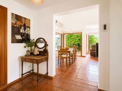 The bright entryway features traditional terracotta flooring and a vintage console table with mirror, opening into a spacious dining area with large windows overlooking lush garden views.