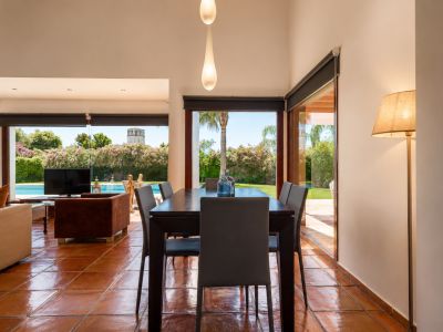 The dining area features a dark wooden table and terracotta floors, illuminated by natural light from large glass doors. It offers direct views of the lush garden, swimming pool, and a distant tower.