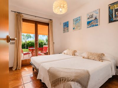 Twin bedroom featuring terracotta floors and direct access to a private outdoor terrace. The large glass door reveals a garden with a palm tree and red seating.