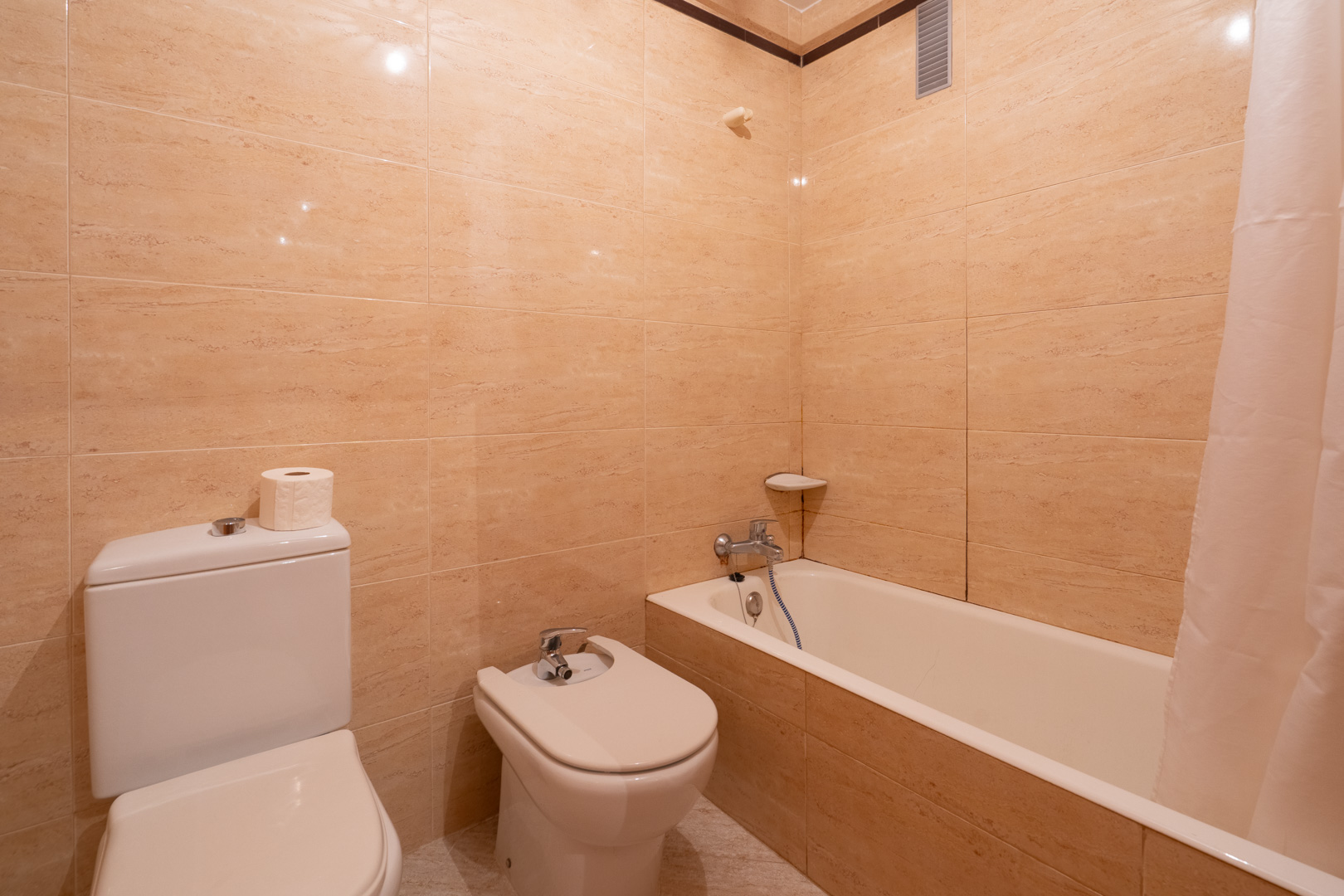 Bathroom featuring beige tiled walls, a white toilet, bidet, and bathtub with a shower curtain. Chrome fixtures and a corner shelf are visible.