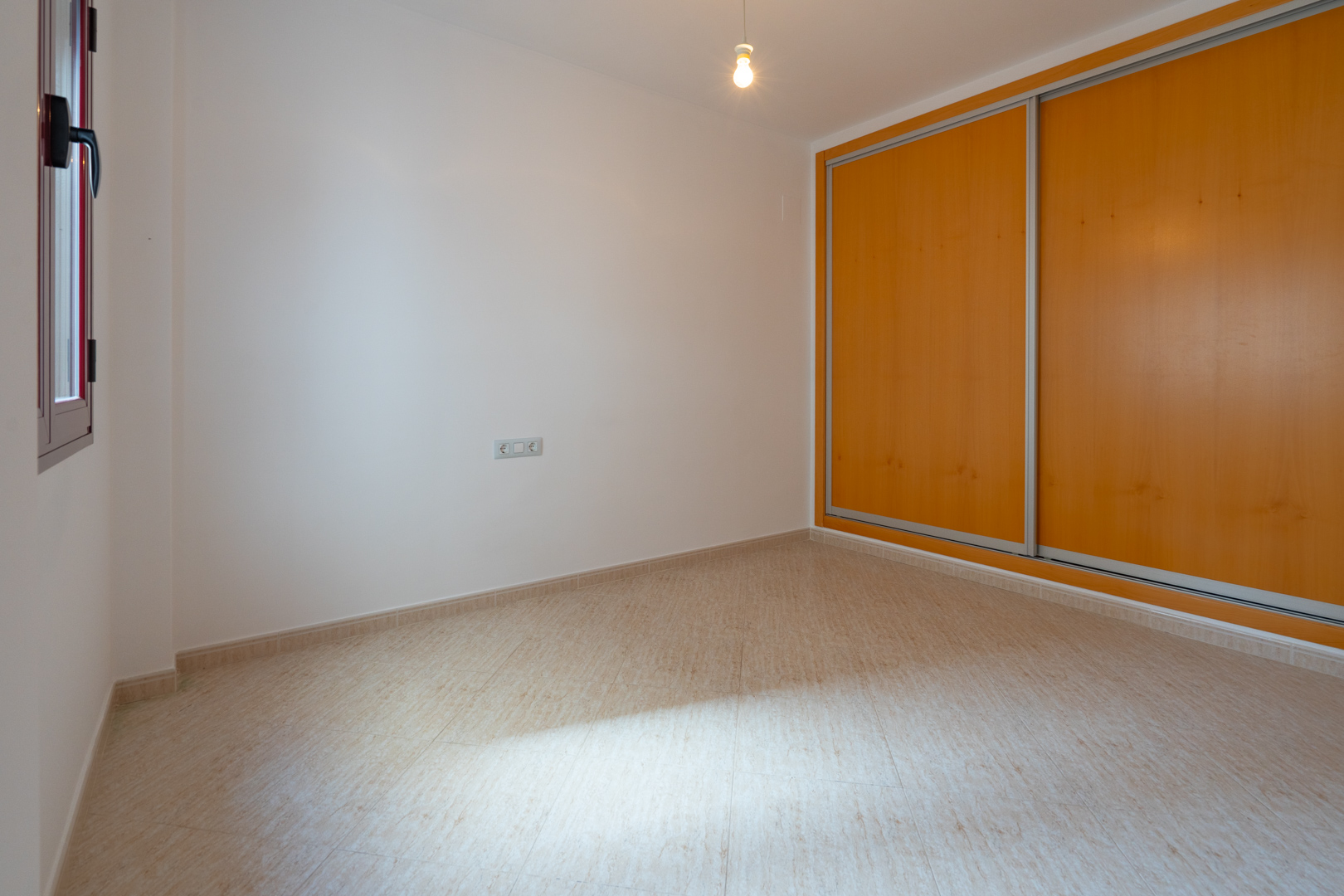 Bright room featuring a built-in wardrobe with two light wood sliding doors and a beige tiled floor. A window with a dark frame provides natural light.