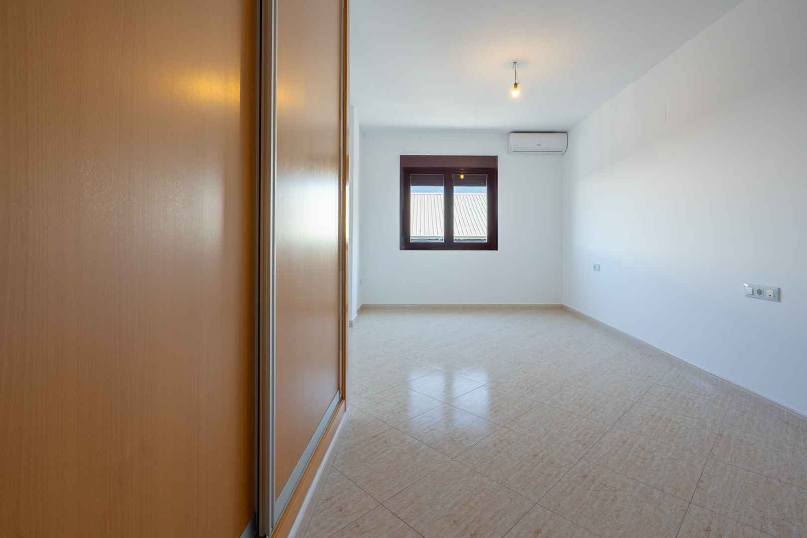 Empty room featuring a light wood built-in wardrobe, diagonally laid beige tiled floor, and a window with a corrugated roof view. Air conditioning unit visible.