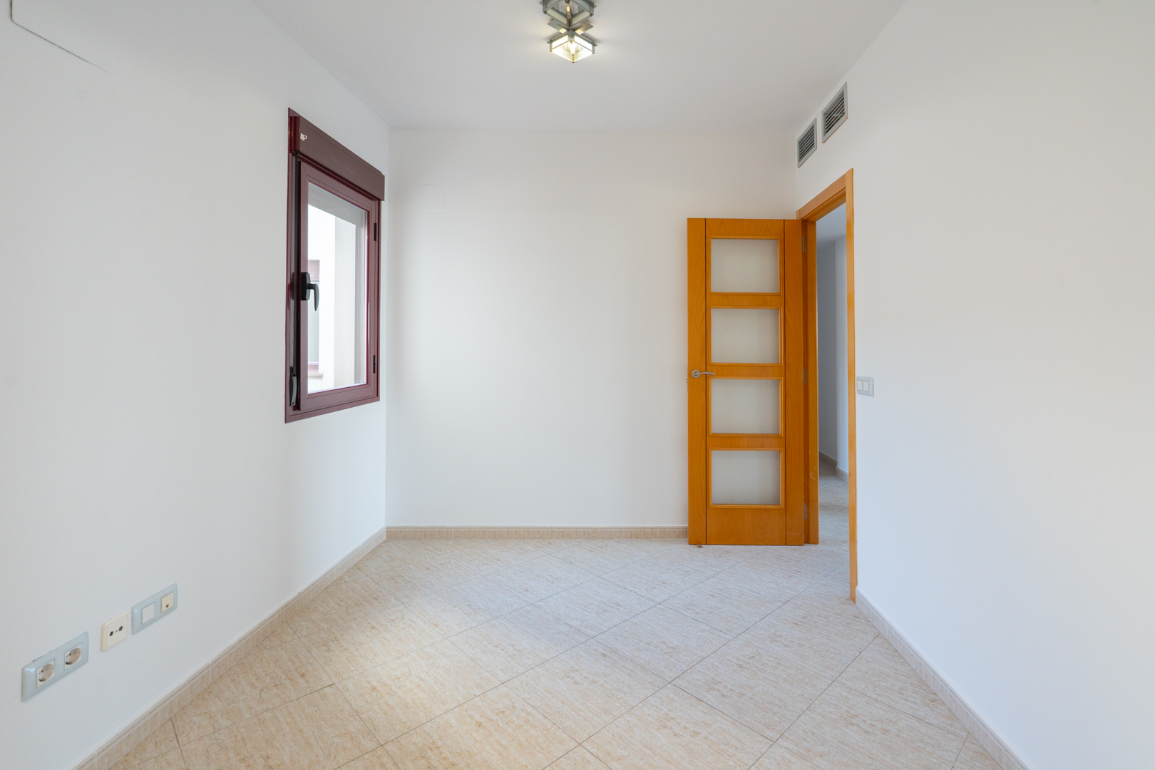 Empty room featuring white walls, a dark red-framed window, and light beige diagonal floor tiles. A wooden door with glass panels and AC vents are visible.