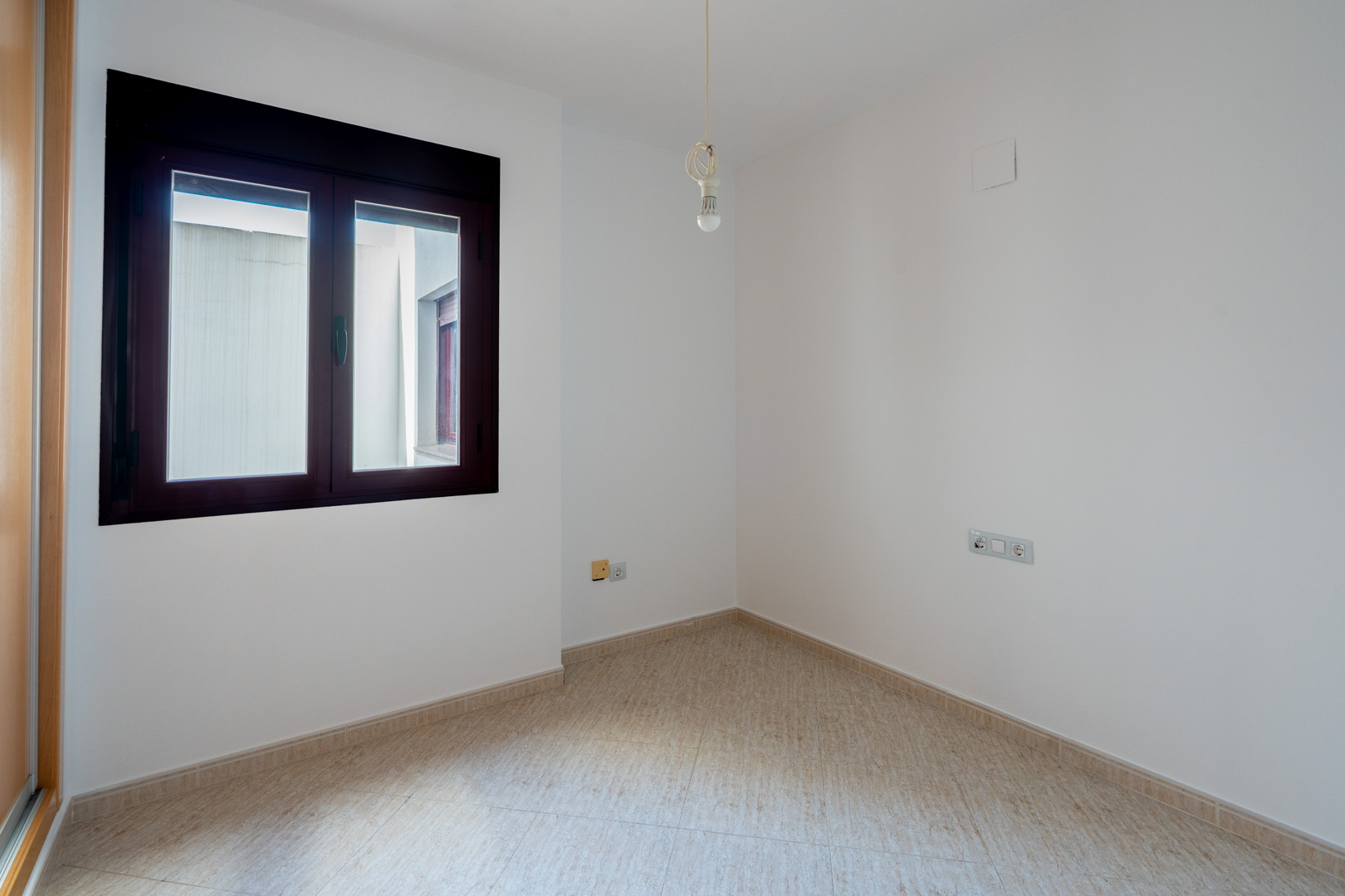 Empty room featuring white walls, light tiled flooring, and a dark-framed window overlooking an interior patio. A single bare bulb light fixture hangs from the ceiling.