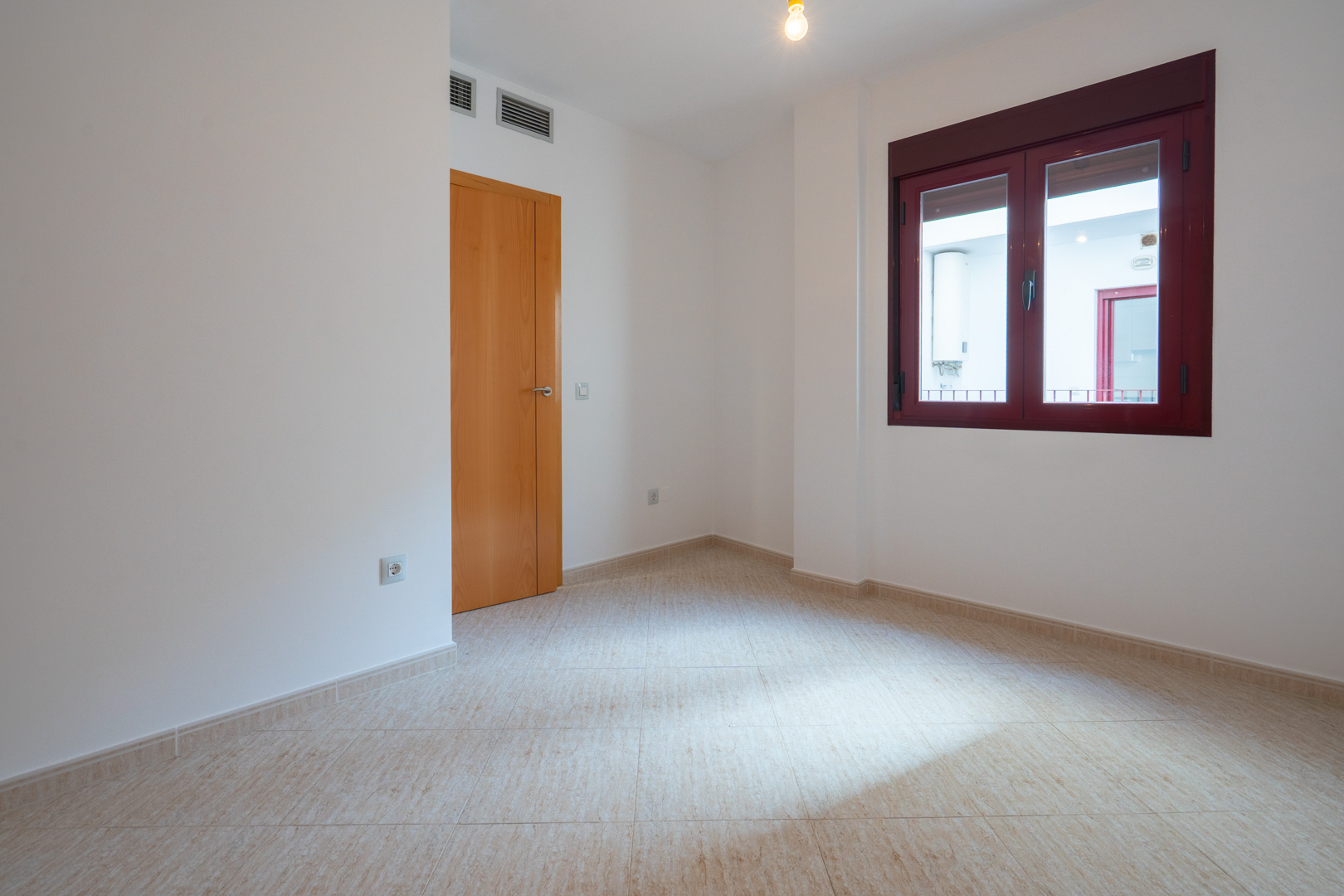 Empty room with light tiled floor and white walls. Features a wooden door and a dark red-framed window, looking out to an interior patio with a water heater. AC vents are visible above the door.