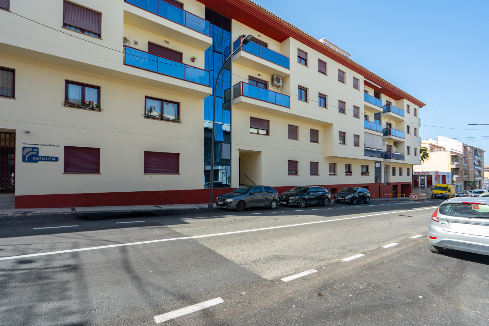 Exterior view of the residential building featuring a light facade, blue glass balconies, windows with maroon shutters, and a vertical glass panel.