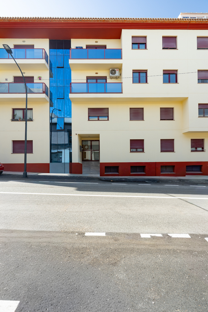 Exterior view of the residential building, featuring cream facade, blue glass balconies, and red roller shutters. A prominent central blue glass section and a street-level entrance with a ramp are visible.