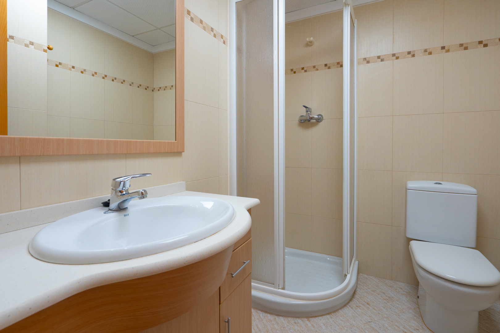 Functional bathroom featuring an oval sink on a light wood vanity, framed mirror, corner shower, and beige tiles with a decorative border.