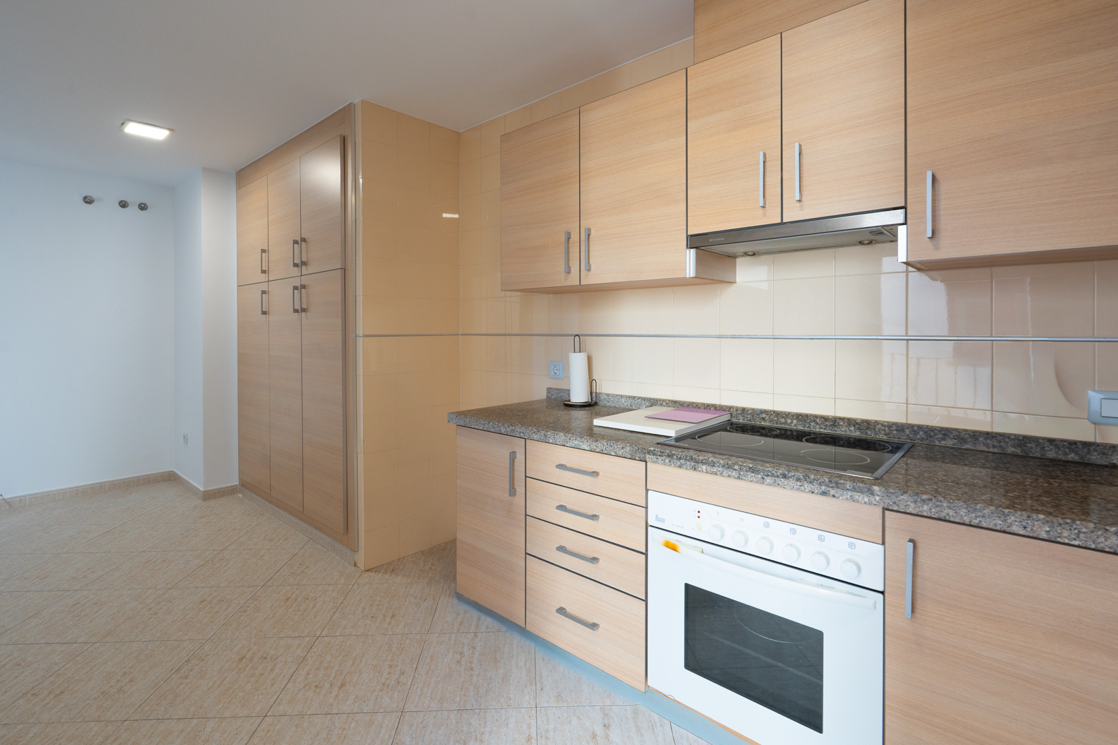 Functional kitchen featuring light wood-effect cabinetry, granite-effect countertop, integrated ceramic hob, and white oven. Tall pantry unit and beige floor tiles are visible.