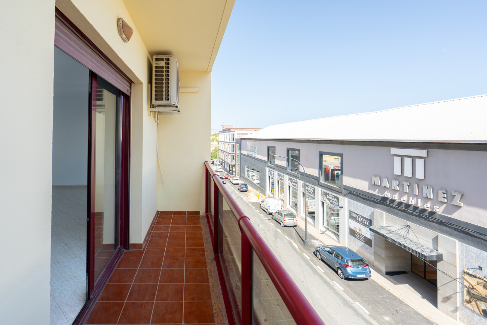 Outdoor balcony featuring terracotta floor tiles and a red railing. An air conditioning unit is visible, offering views of the street and "Martinez Muebles" store.