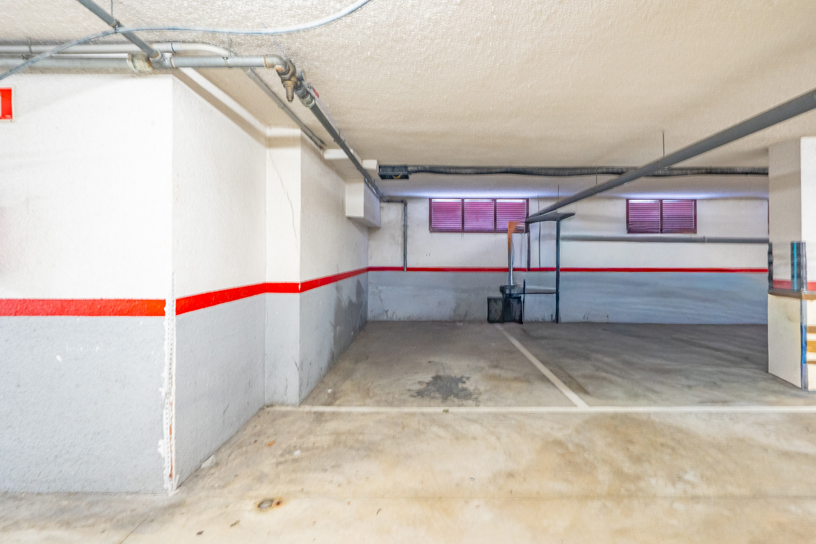 Private underground parking space with concrete floor, white and grey walls featuring a red stripe, and exposed ceiling pipes. Two small windows with red blinds are visible.