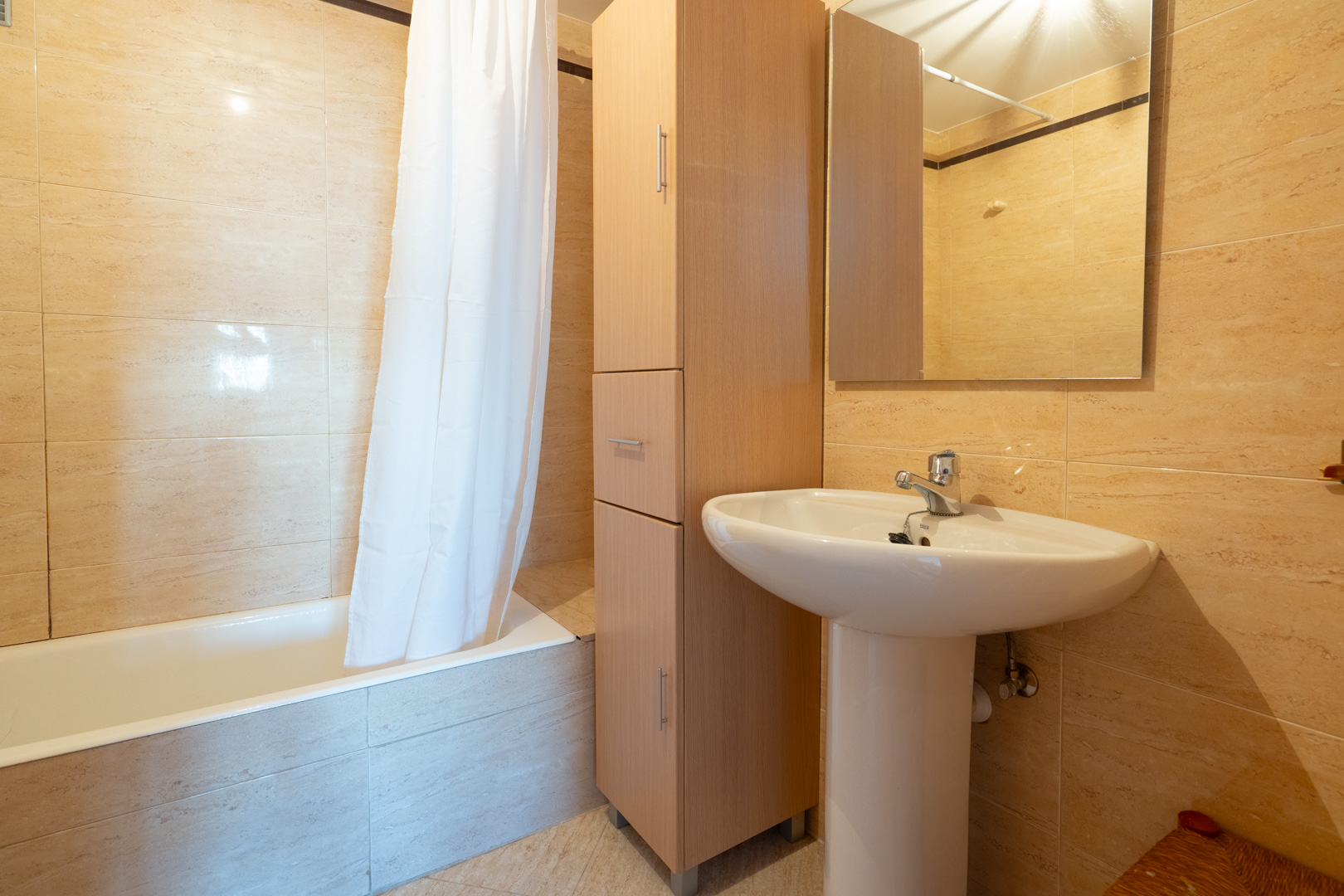 Second bathroom featuring a white pedestal sink, a tall wooden storage cabinet, and a bathtub with a shower curtain. Walls are finished with light beige tiles.