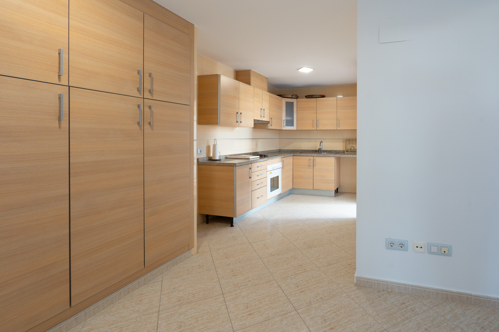 Spacious kitchen featuring light wood cabinetry, speckled granite countertops, and an integrated oven. A large matching pantry unit stands on the left, complementing the beige tiled floor.