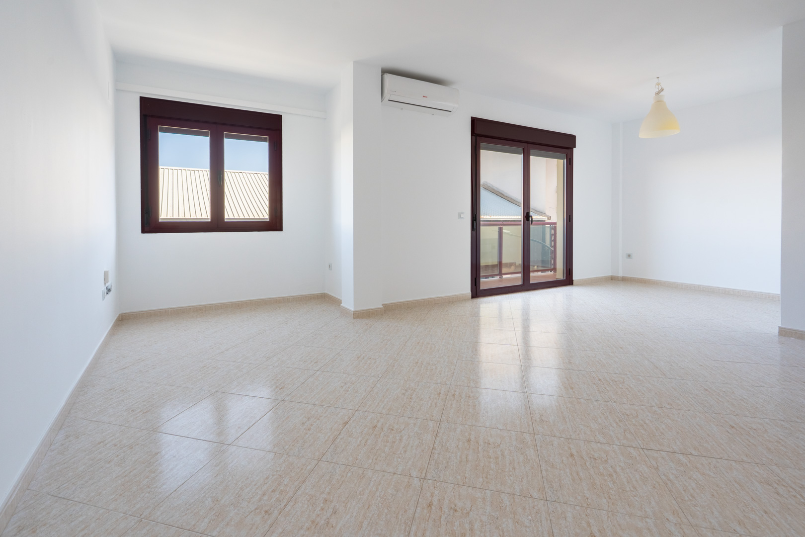 Spacious room featuring light beige tiled flooring, white walls, a window, and a sliding door with dark brown frames leading to a terrace. Includes a wall-mounted AC unit.