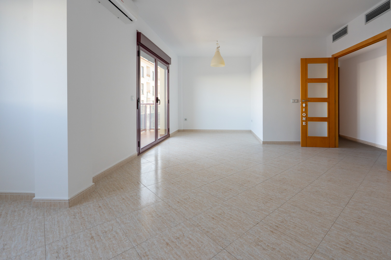 Spacious room with light beige ceramic tiled flooring. Features a large sliding glass door with a dark red frame to a terrace, a split AC unit, and a light wooden door with frosted glass panels.