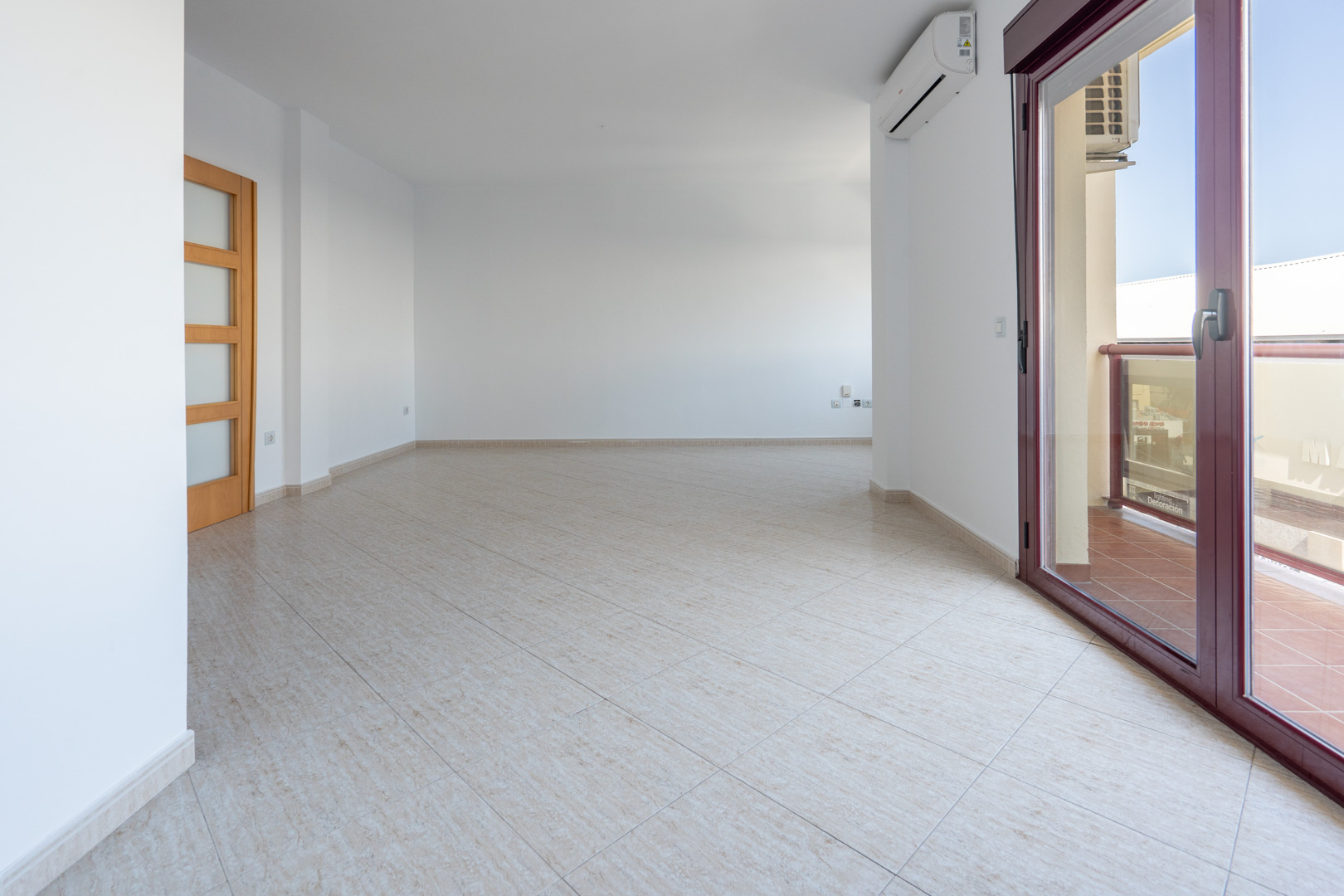 Spacious, unfurnished living-dining area with light tiled flooring, a wooden door with frosted glass panels, and access to a terrace via a dark red aluminum sliding door. An AC unit is visible.