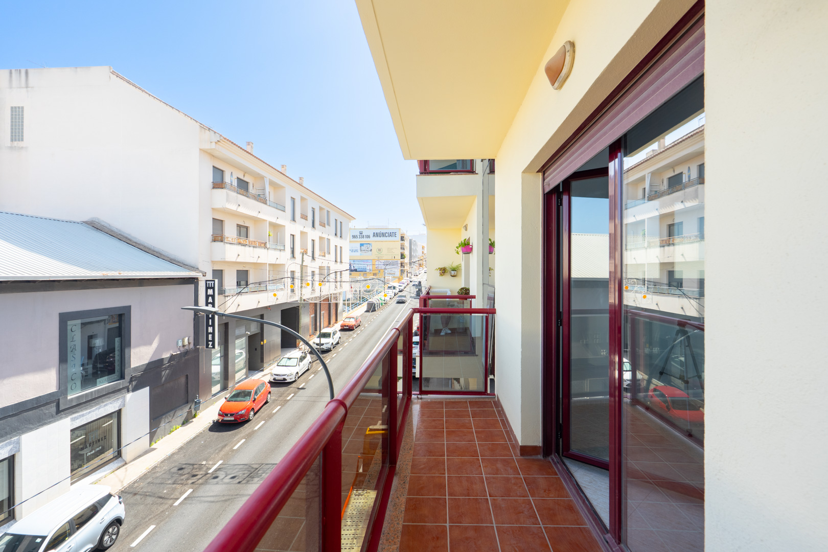 Sunny balcony featuring terracotta floor tiles and a red-framed glass railing, overlooking an urban street with buildings and traffic.