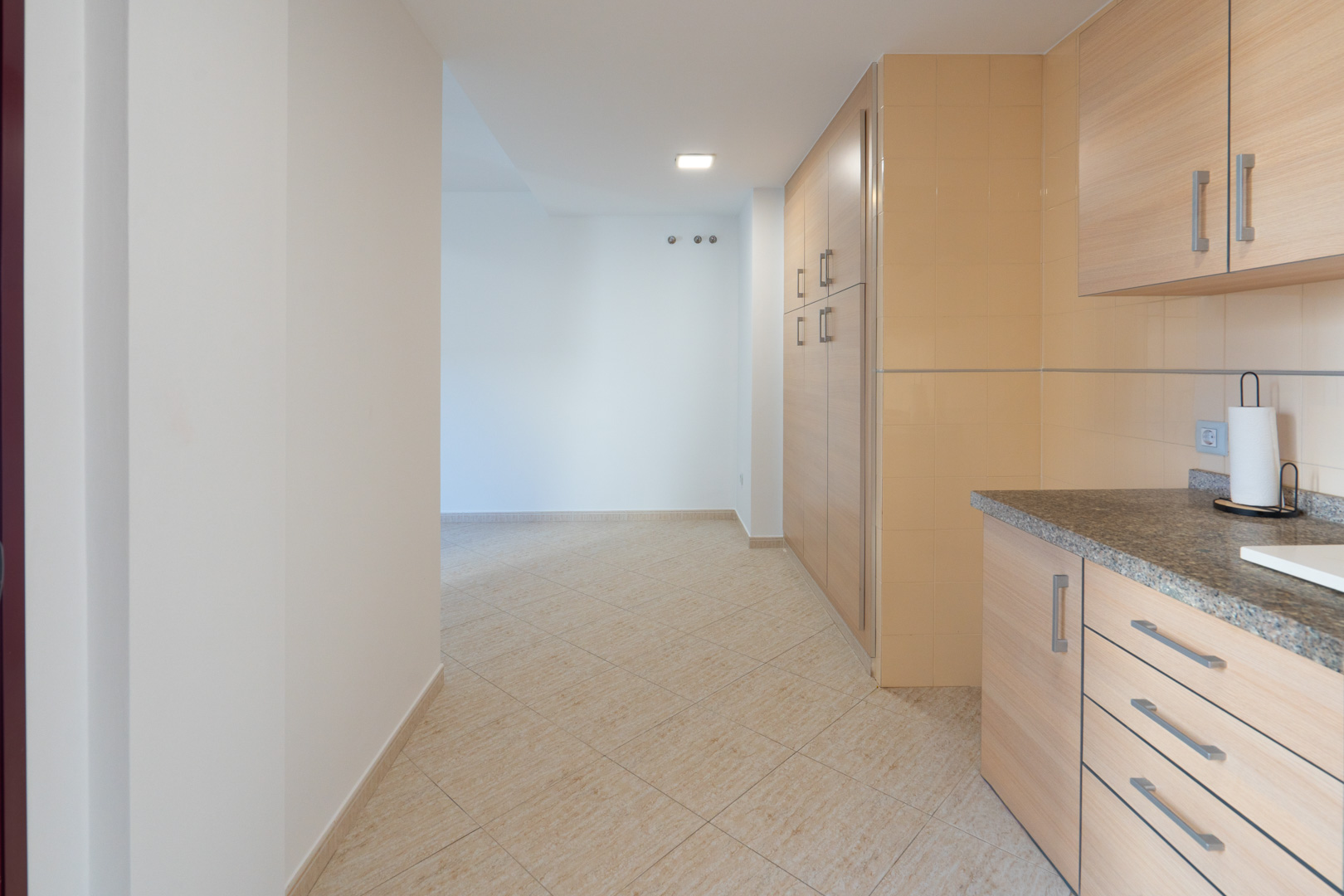 Transition from the kitchen, featuring granite countertop and light wood cabinets, to a hallway with beige ceramic floor and wall hooks.