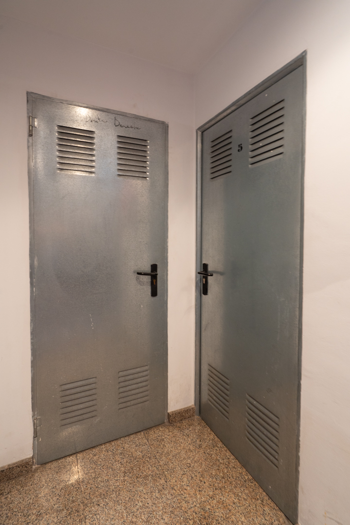 Two grey metallic doors with upper and lower vents, one marked 'Benissa Denia' and the other with the number '5', set on a speckled granite floor.
