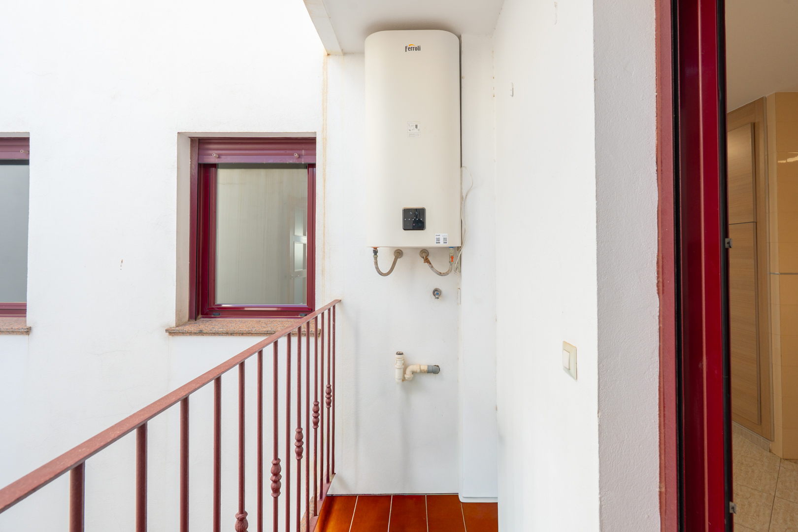 Utility patio featuring a wall-mounted Ferroli water heater, terracotta floor tiles, and a red metal railing. A burgundy-framed window with a granite sill and a matching door frame are visible.
