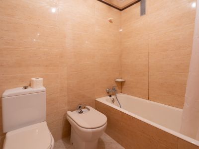Bathroom featuring beige tiled walls, a white toilet, bidet, and bathtub with a shower curtain. Chrome fixtures and a corner shelf are visible.