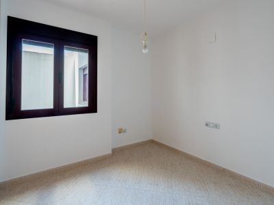 Empty room featuring white walls, light tiled flooring, and a dark-framed window overlooking an interior patio. A single bare bulb light fixture hangs from the ceiling.